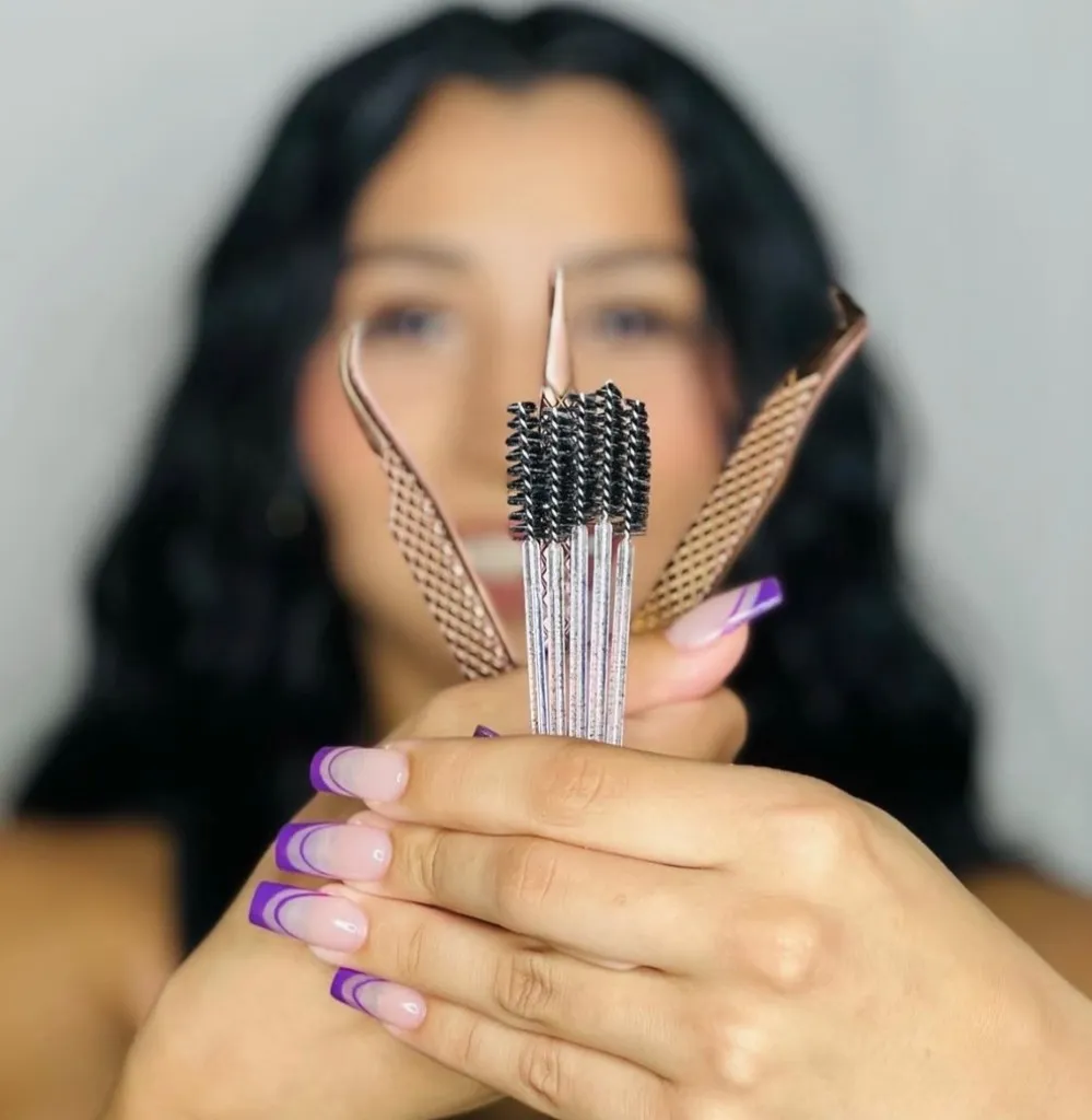 Gigi holding rose-gold lash tweezers and spoolie brushes, with her long dark hair softly blurred in the background